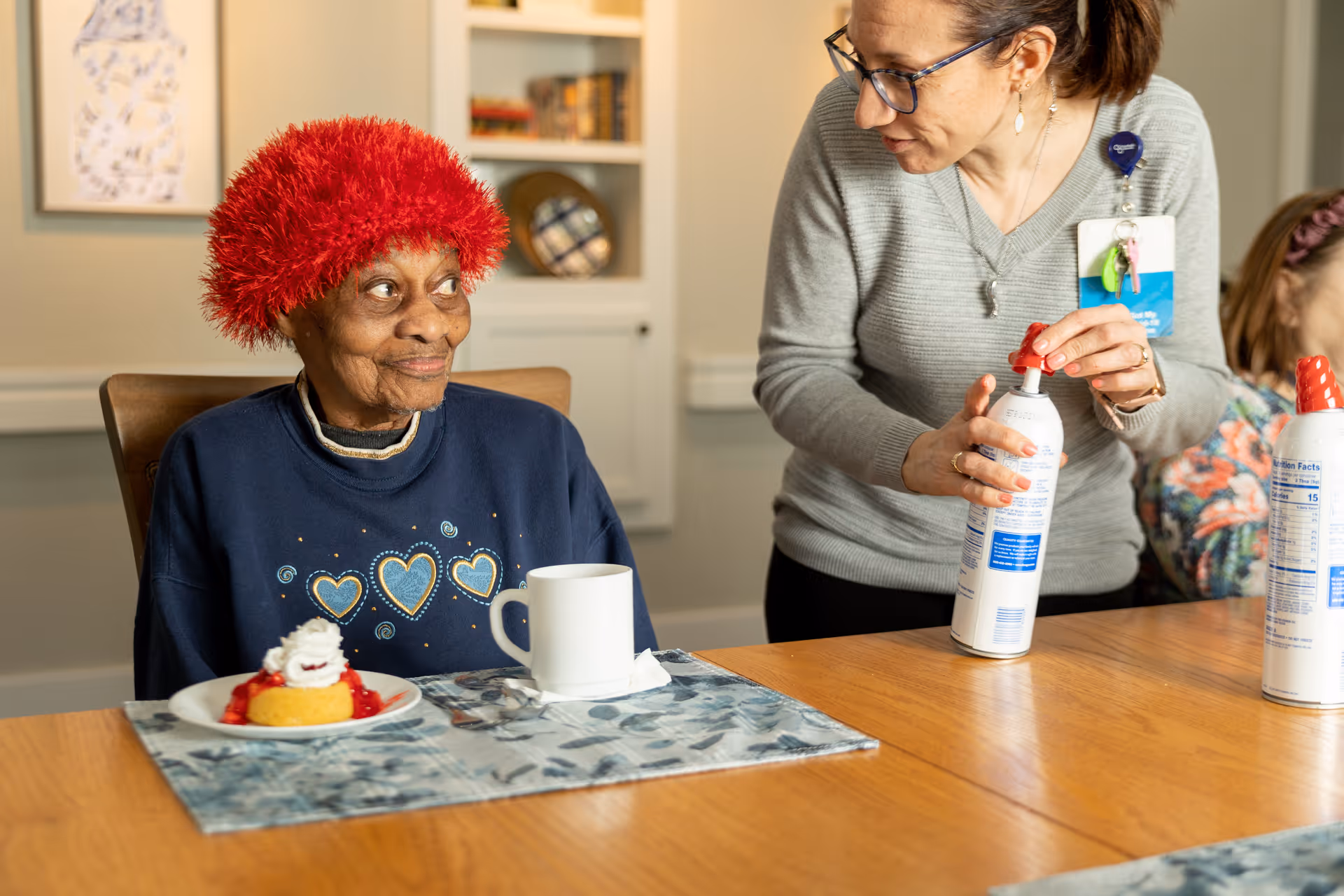 An elderly woman wearing a red fuzzy hat smiles at a caregiver who is opening a can of whipped cream across a dining table with a dessert and mug.