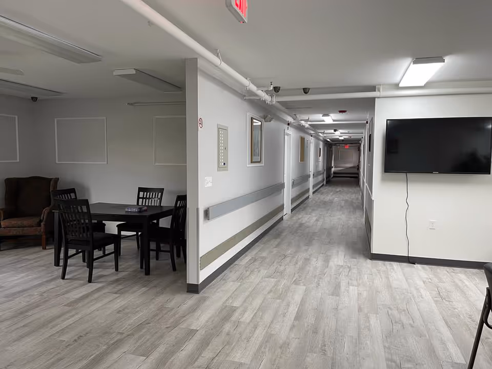 Interior hallway and common seating area with a table and chairs, wall-mounted TV, and long corridor with handrails.