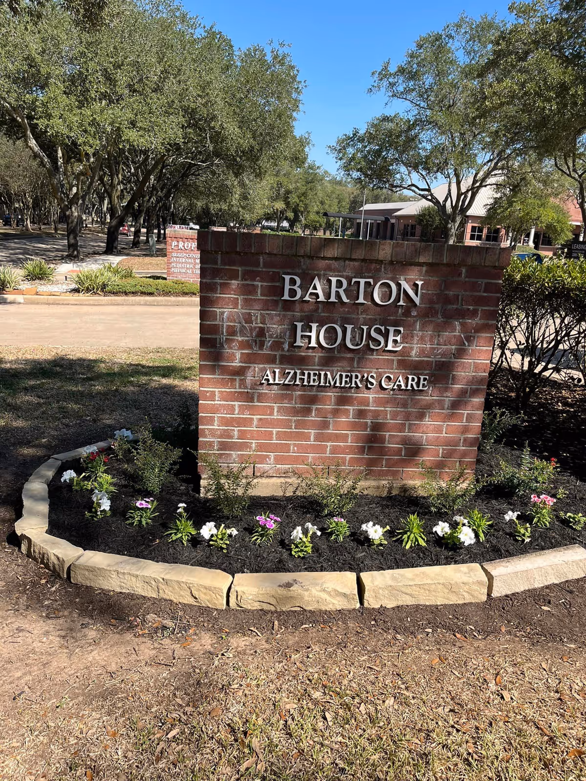 Brick entrance sign reading 'Barton House Alzheimer's Care' surrounded by a flowerbed with trees and a building in the background.