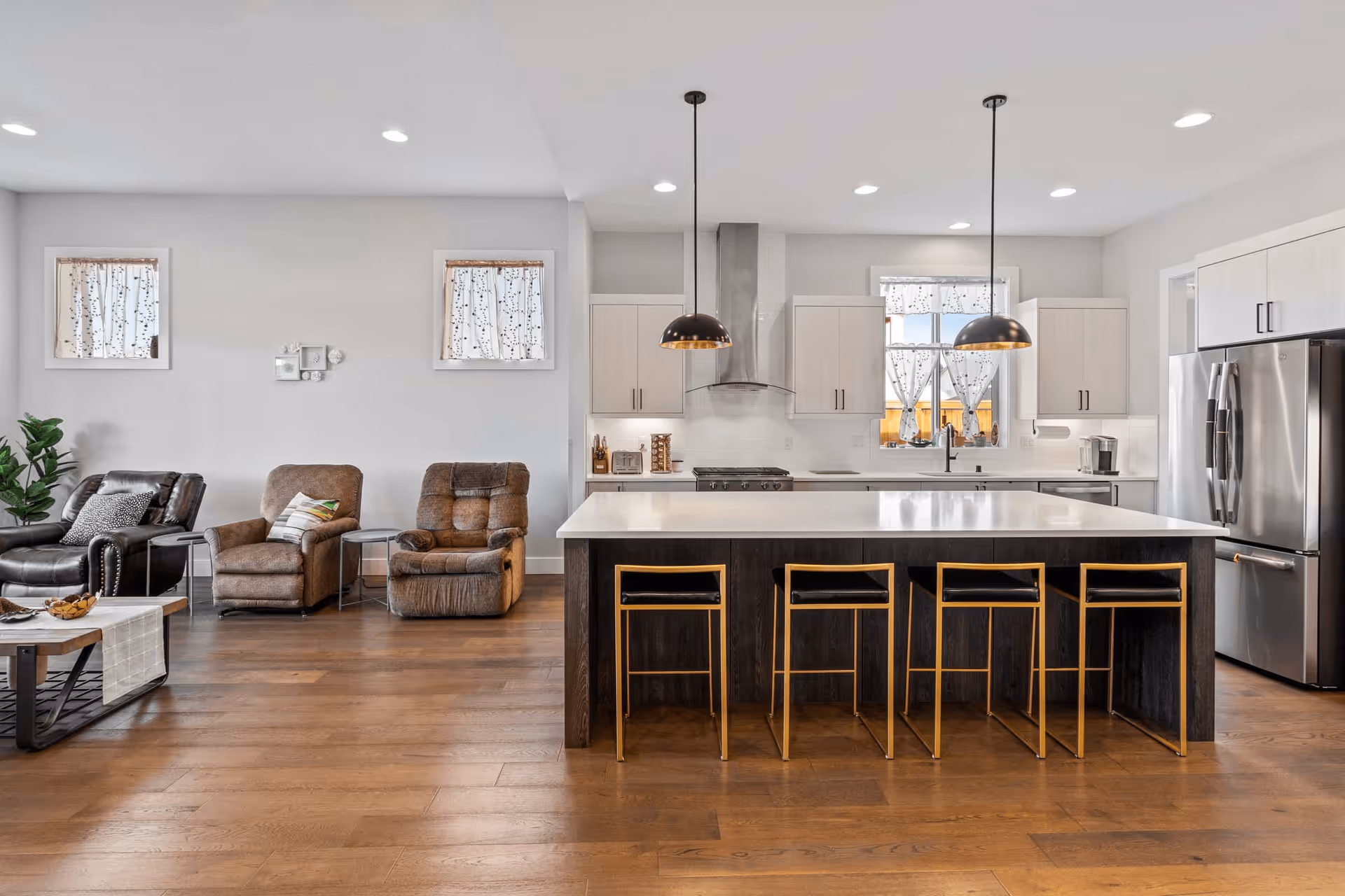 A modern open-concept living area featuring a kitchen with a large island and four bar stools. The kitchen has white cabinetry, stainless steel appliances, two pendant lights hanging over the island, and a window with patterned curtains. Adjacent to the kitchen is a seating area with three armchairs and a coffee table on wooden flooring.