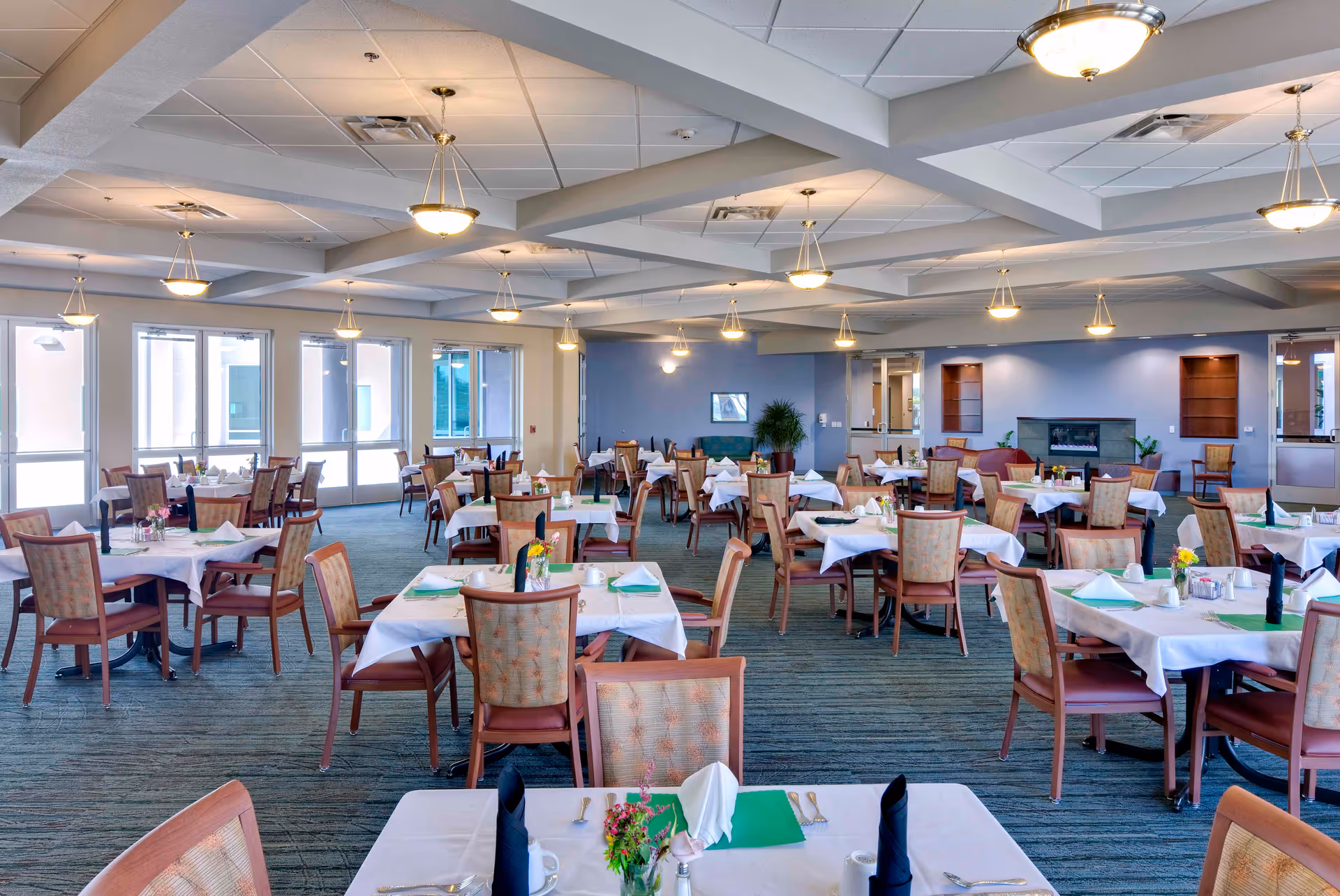 A spacious dining room in a senior living facility with multiple tables covered in white tablecloths, each set with napkins, utensils, and small flower arrangements. The room has large windows allowing natural light, a carpeted floor, and ceiling lights evenly distributed across the ceiling.