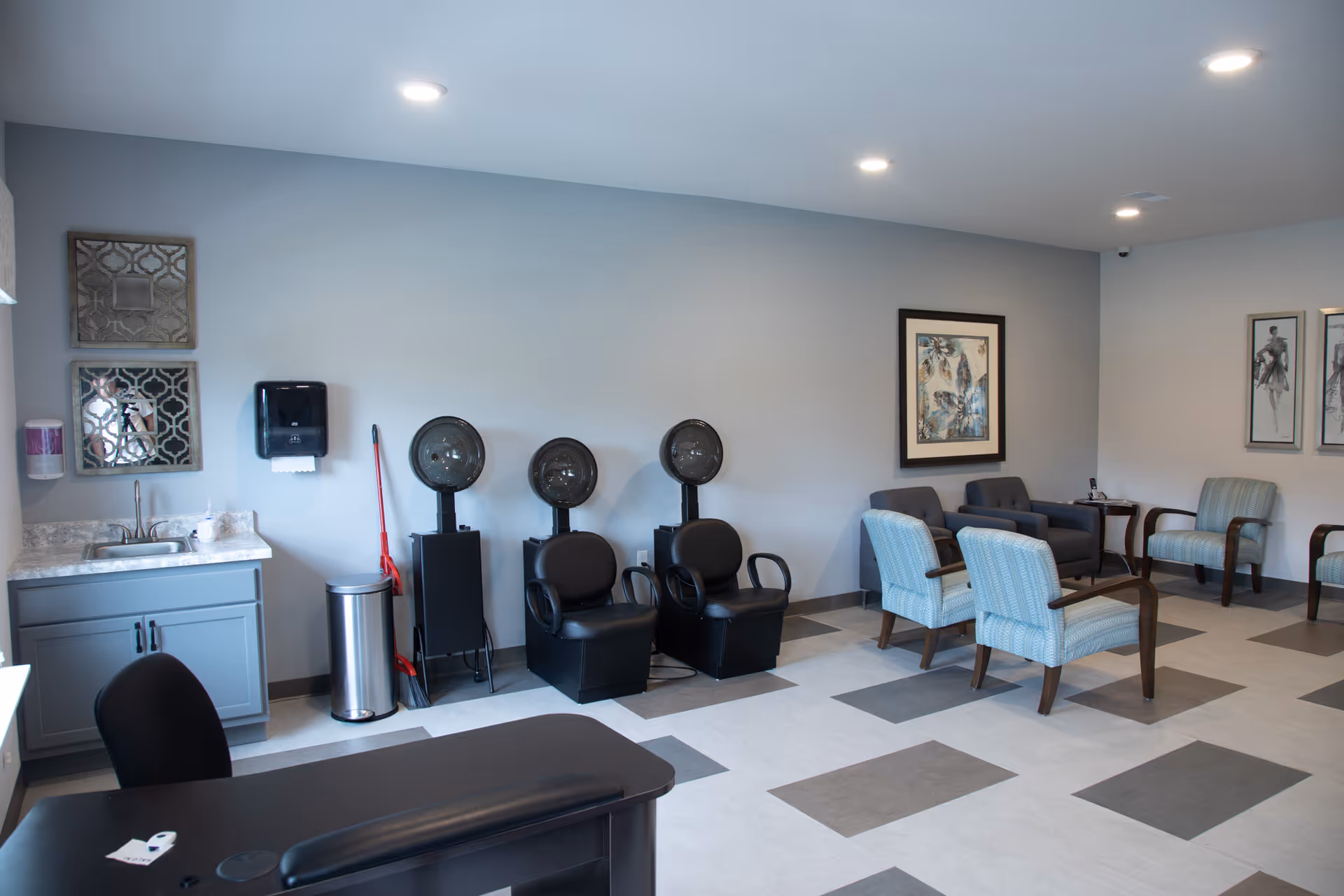 Interior of a salon area in a senior living facility with two black hair dryer chairs, a small sink with a cabinet, a trash can, a mop, and several chairs arranged around the room. The walls are decorated with framed artwork.