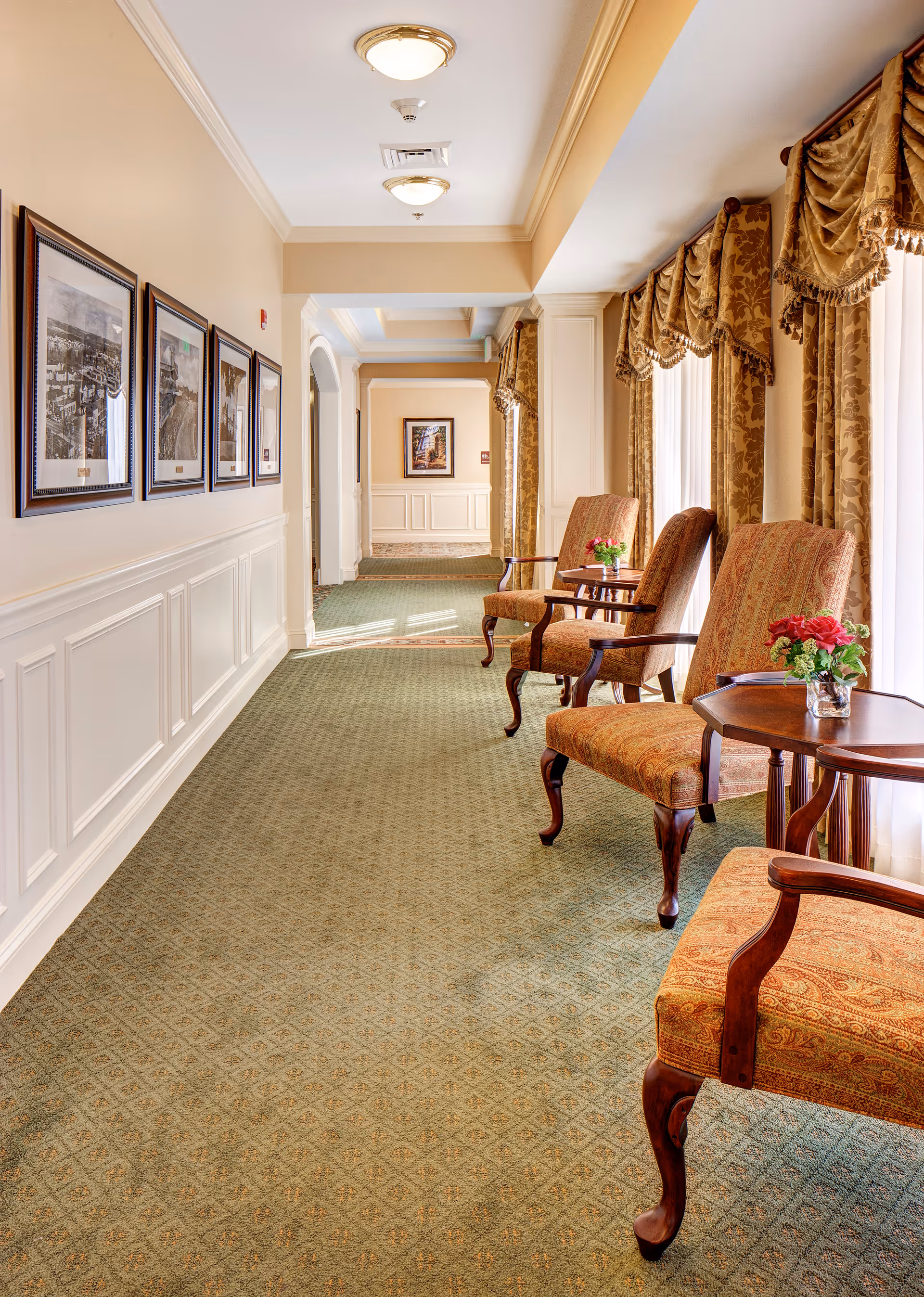 A well-lit hallway in a senior living facility with patterned green carpet, beige walls with white wainscoting, and framed black-and-white photographs on the left wall. On the right side, there are three upholstered armchairs with wooden legs and arms, each paired with a small wooden side table holding a small vase with red flowers. Large windows with ornate gold curtains allow natural light to fill the space.