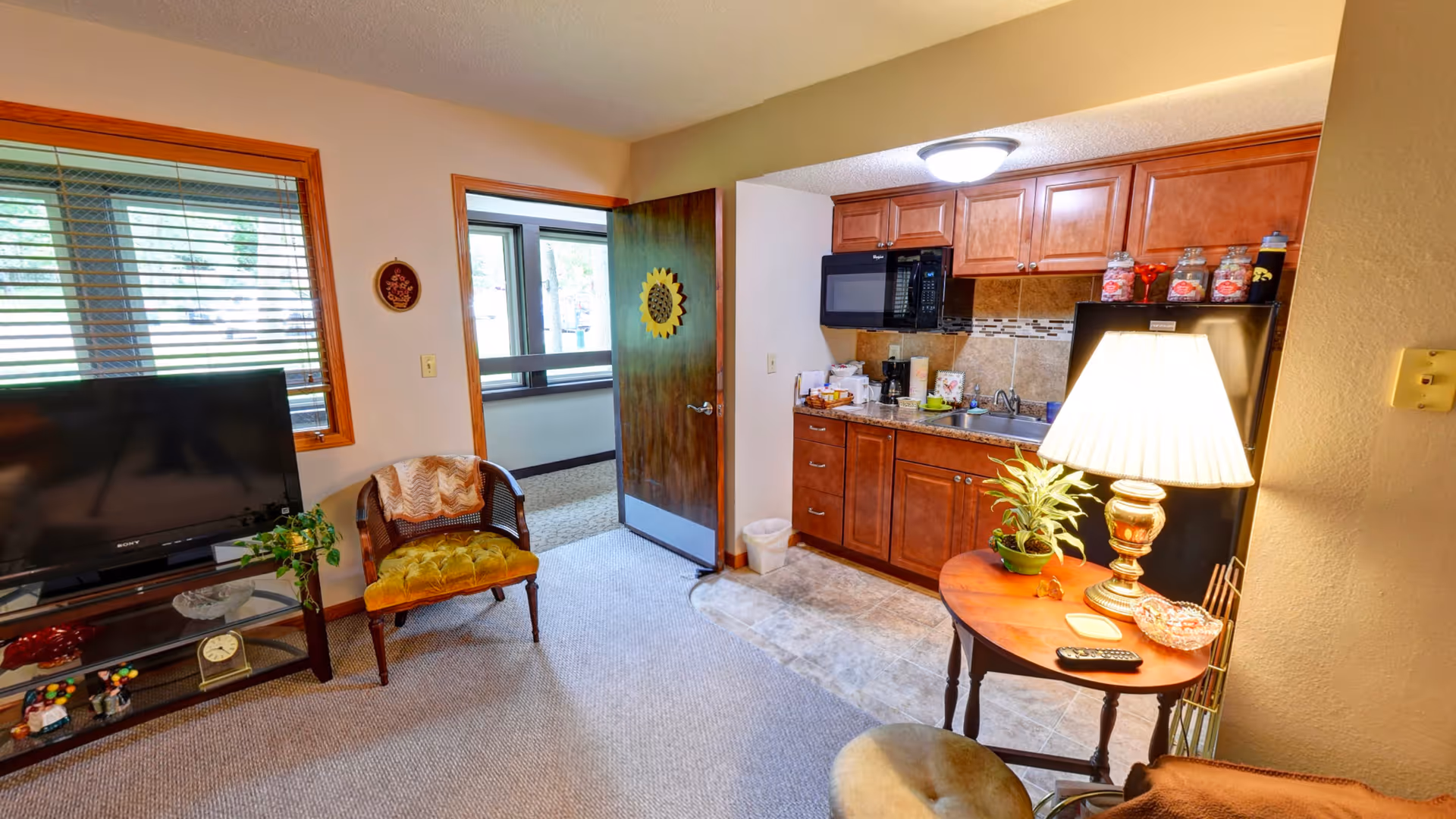 Interior view of a senior living facility room at Garnett Place featuring a small kitchenette with wooden cabinets, a microwave, and a refrigerator. Adjacent to the kitchenette is a round wooden table with a lamp, a plant, and a remote control. There is a green cushioned chair next to a window with wooden blinds, and a flat-screen TV on a stand with decorative items below. An open door leads to another room with large windows.