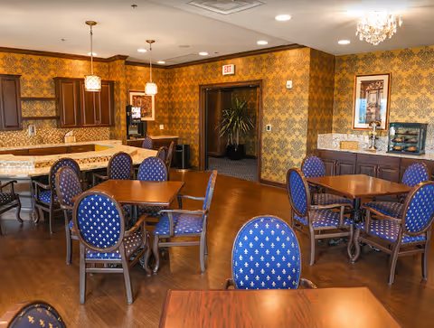 A dining area with several wooden tables and blue upholstered chairs with a fleur-de-lis pattern. The room has ornate yellow and brown wallpaper, wooden flooring, and a kitchen area with granite countertops and wooden cabinets. Pendant lights hang above the kitchen counter, and a chandelier is visible on the ceiling. There is a doorway leading to another room with a plant visible inside.