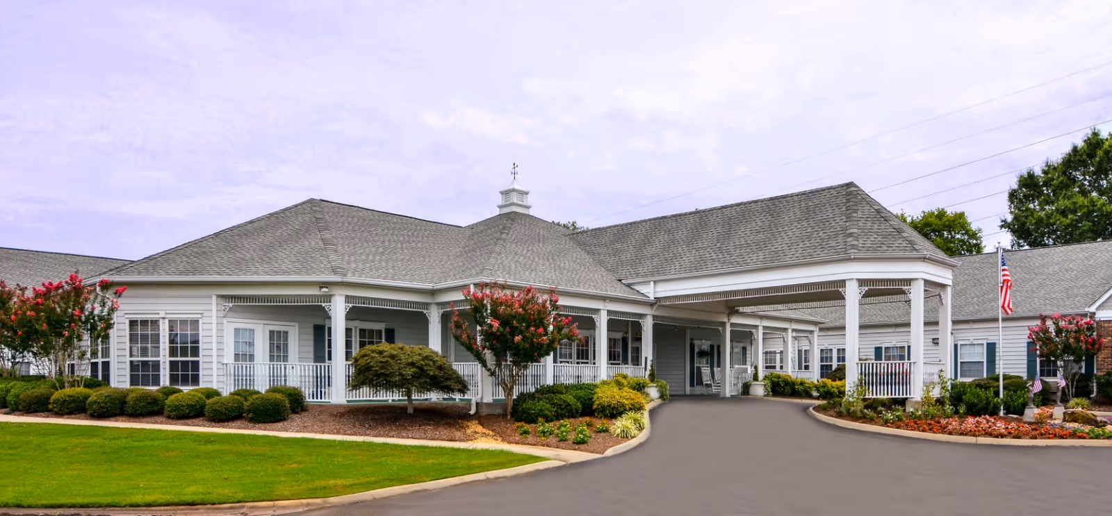 Exterior view of a single-story senior living facility building with a covered entrance, manicured bushes, flowering trees, and an American flag on a flagpole.