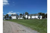 Wide view of a large white multi-building facility with multiple windows and pitched roofs, situated on a grassy lawn under a blue sky with scattered clouds. A dirt road runs alongside the lawn leading towards the buildings.