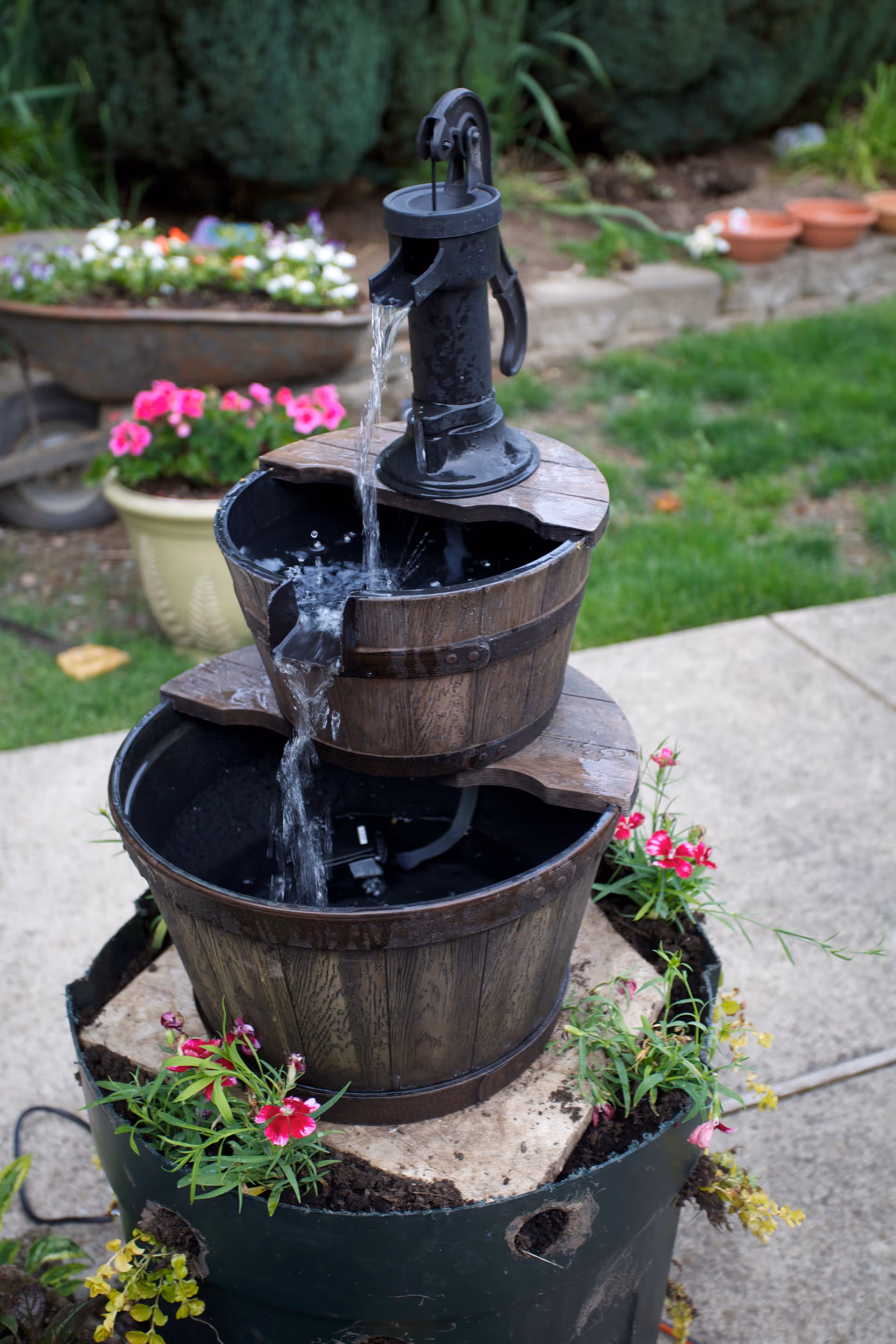 A decorative outdoor water fountain designed to look like two stacked wooden barrels with water flowing from a black hand pump at the top barrel into the lower barrel. The fountain is surrounded by small flowering plants and is placed on a concrete surface with grass and garden elements in the background.