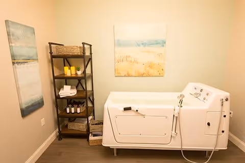 Small bathing room with a walk-in tub, a shelving unit holding towels and toiletries, and coastal-themed wall art.