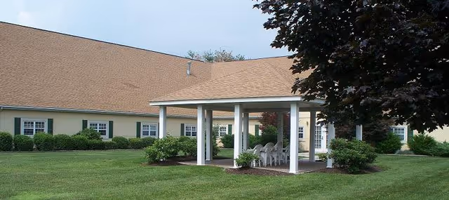 Single-story building with beige siding and green shutters surrounding a grassy courtyard with a white gazebo and several white chairs underneath. Trees and shrubs are planted around the building and gazebo.