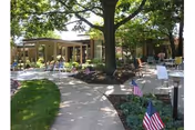 Shaded outdoor courtyard with a paved walkway, trees, seating areas, and small American flags.