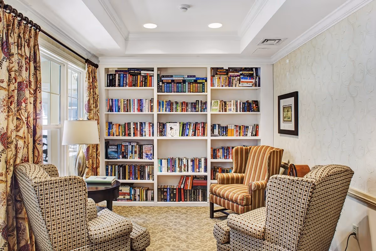 A cozy reading room with three upholstered armchairs arranged around a small round wooden table with a lamp. Behind the chairs is a large white bookshelf filled with various books. The room has patterned curtains on the window and light-colored wallpaper with a subtle design.