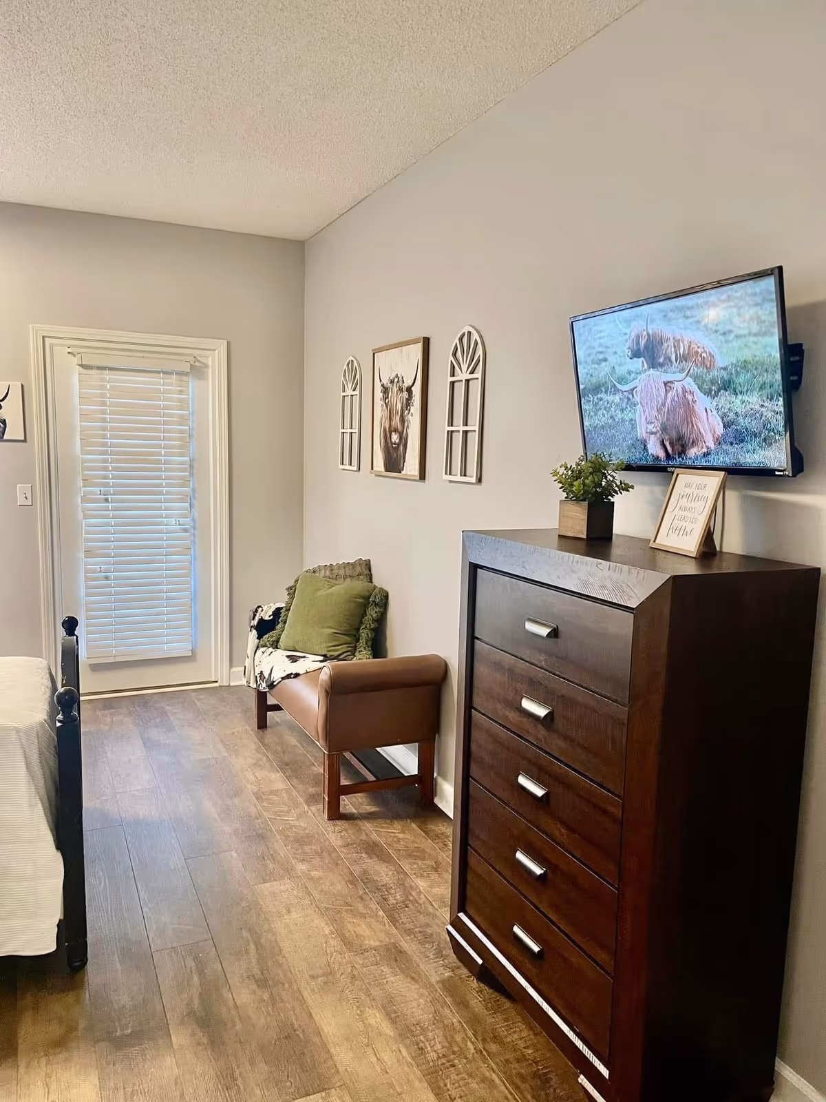 A senior living bedroom featuring a wooden floor, a dark wood chest of drawers with a small plant and framed quote on top, a wall-mounted TV displaying an image of two highland cows, a brown armchair with green and patterned cushions, and three decorative wall hangings including a framed picture of a highland cow and two window-shaped mirrors. A door with closed blinds is visible at the end of the room.