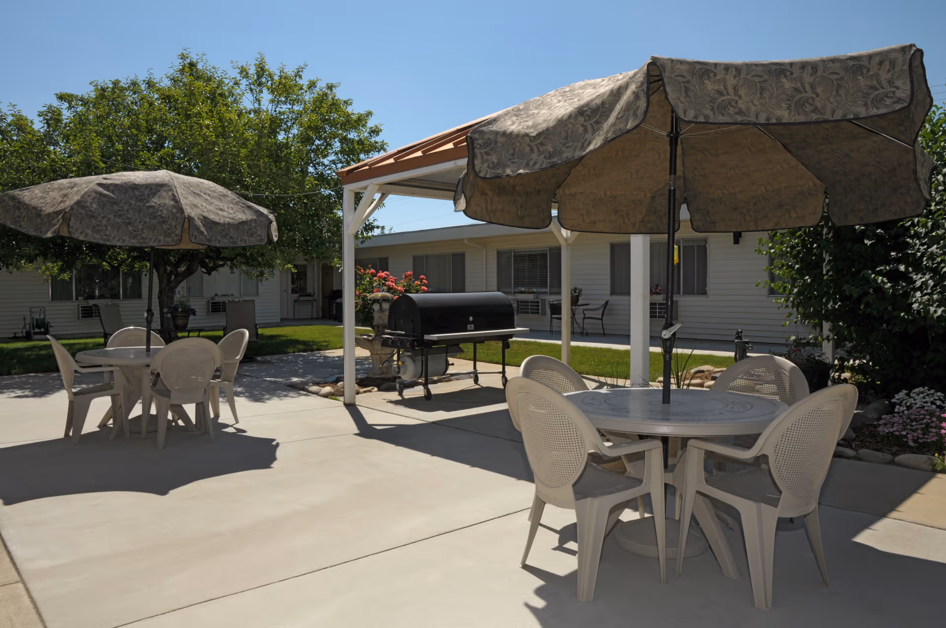 Outdoor patio with round tables and umbrellas, plastic chairs, a barbecue grill, and a single-story building in the background.