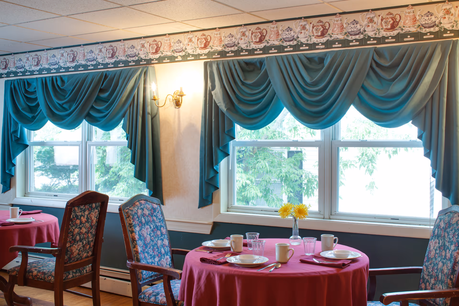 Dining area with round tables covered in red tablecloths, set with plates, cups, glasses, and silverware. The chairs have floral upholstery. Large windows with teal drapes and valances let in natural light, and a small vase with yellow flowers is on one table.