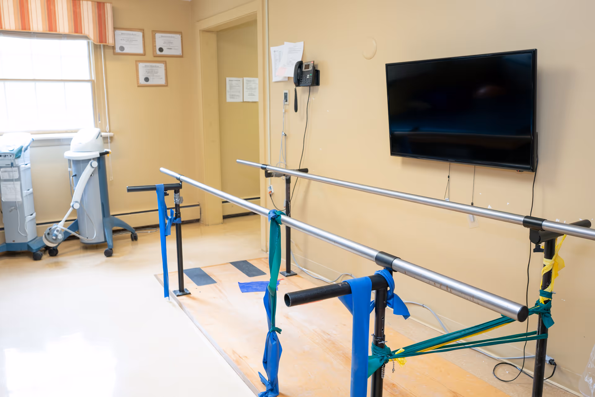 A physical therapy room with parallel bars equipped with resistance bands for walking exercises. The room has beige walls, a mounted flat-screen TV, a telephone on the wall, and medical equipment near a window with striped curtains. Certificates are displayed on the wall near the door.