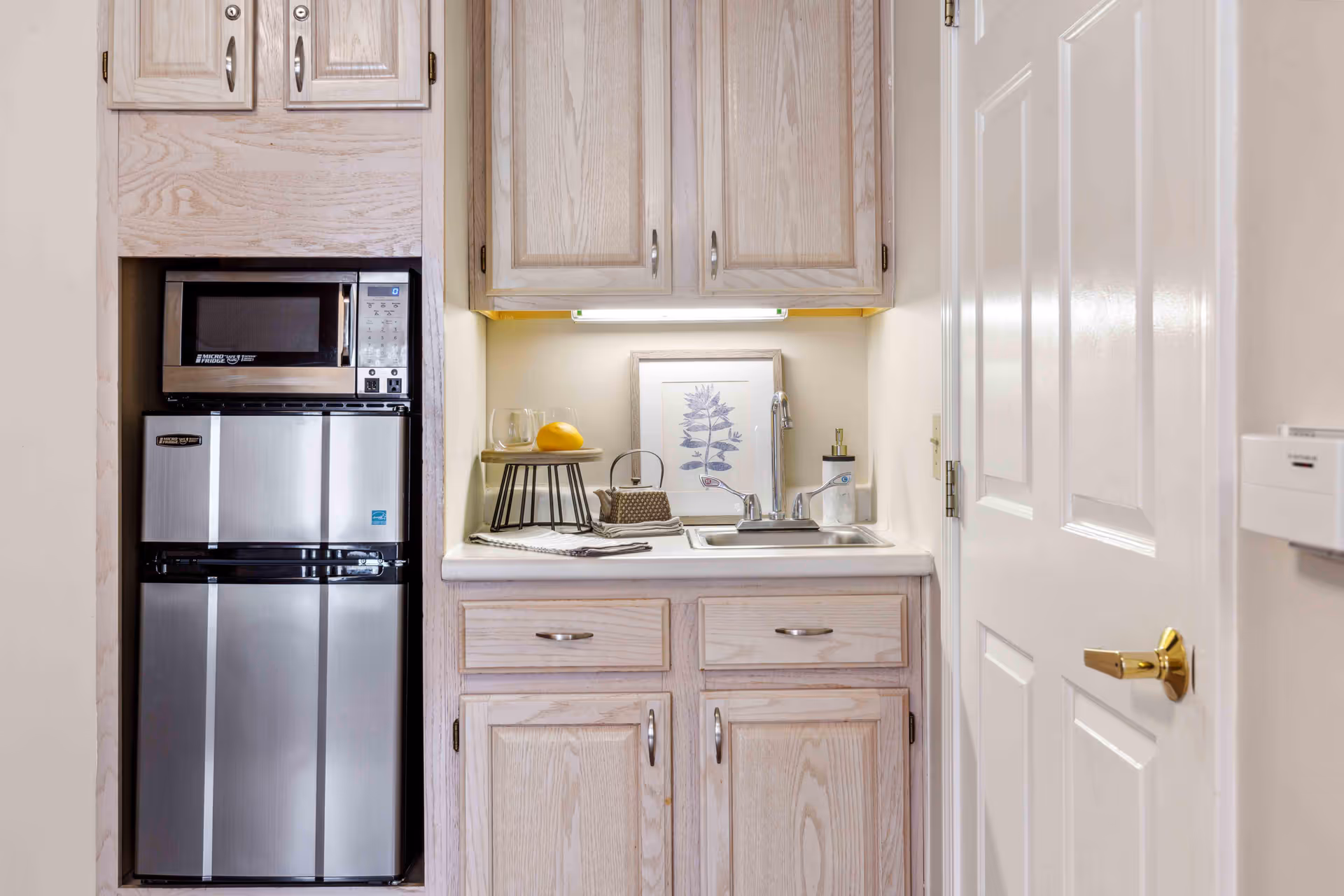 A small kitchenette area with light wood cabinets, a stainless steel microwave and mini refrigerator, a sink with a faucet, and a framed botanical print on the wall. There are also a glass, a lemon, and a small decorative basket on the counter.