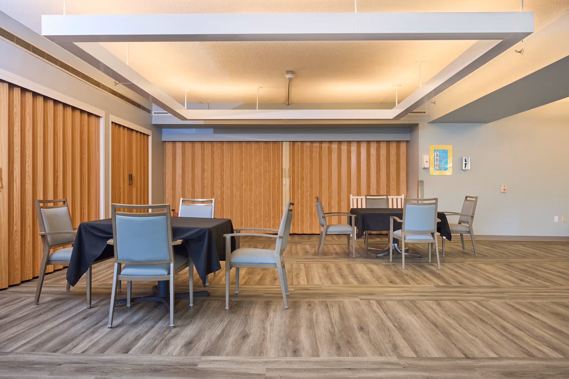 Spacious dining/activity room with several tables draped in black tablecloths and chairs arranged on wood-patterned flooring.