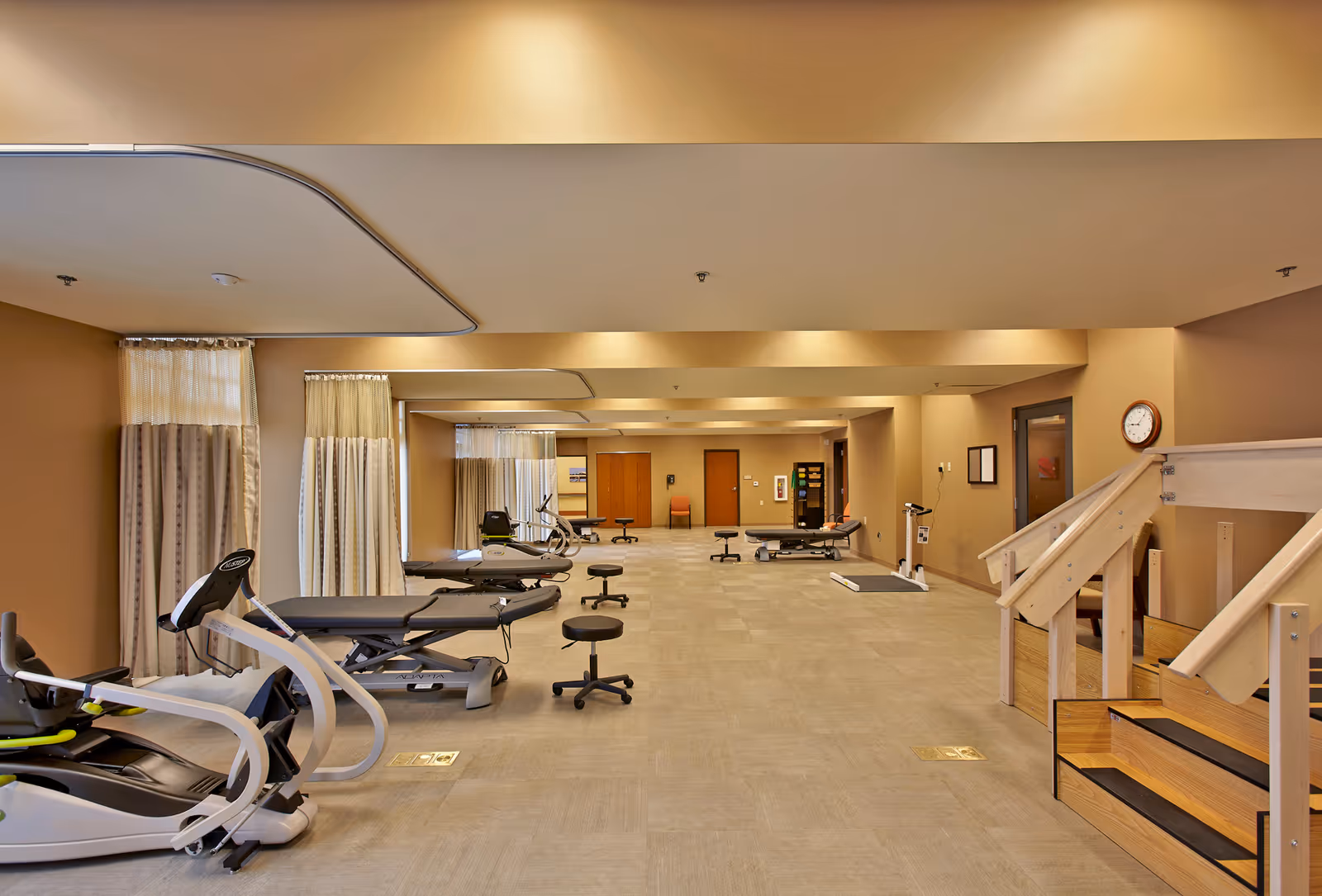 Spacious rehab/exercise room with therapy tables, exercise machines, stools, and a practice staircase in a well-lit interior.