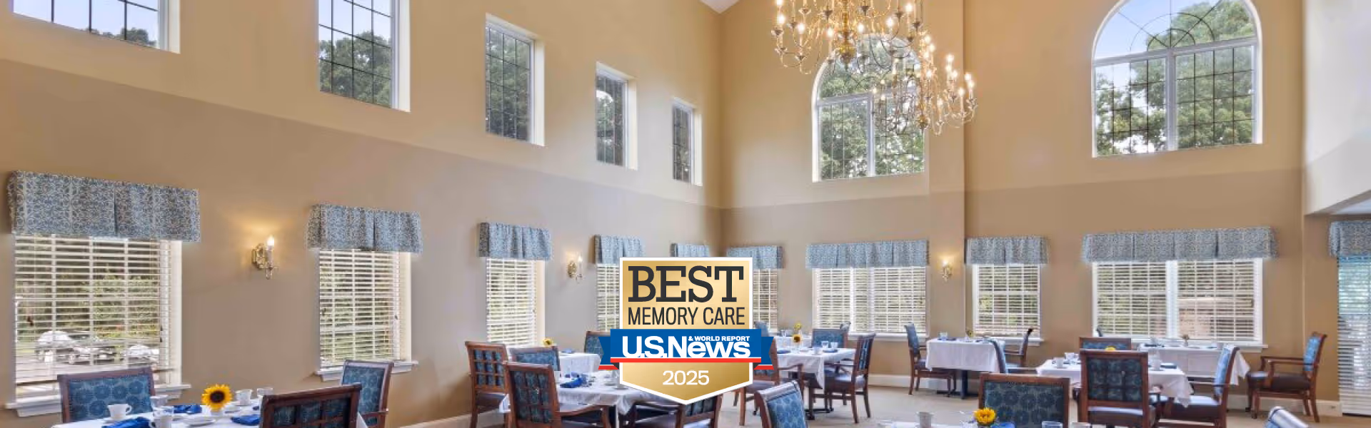 A spacious dining room with high ceilings and large windows letting in natural light. The room features multiple tables covered with white tablecloths, each set with chairs and tableware. Blue patterned valances adorn the windows, and a large chandelier hangs from the ceiling. Outside greenery is visible through the windows.