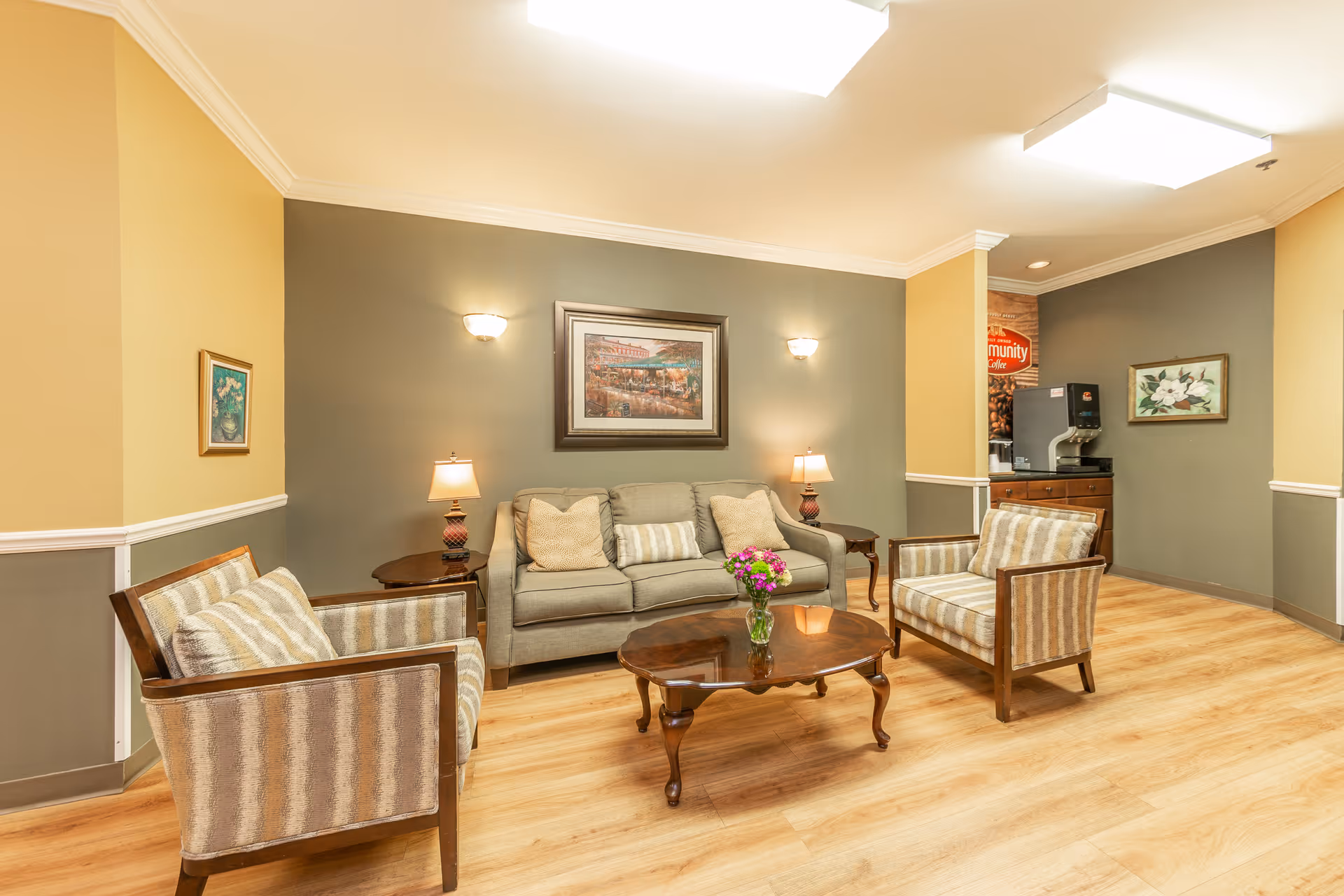 A cozy assisted-living common room with a sofa, two armchairs, wooden coffee table with flowers, lamps, and wall art.