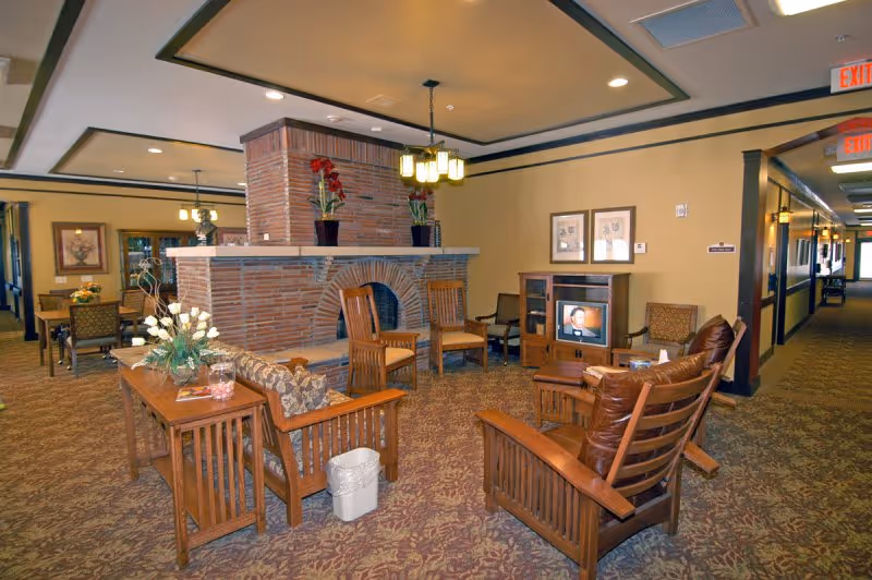 A cozy common area in a retirement center featuring wooden chairs with cushions arranged around a brick fireplace. There is a small TV on a wooden stand, a table with a flower arrangement, and framed pictures on the walls. The space has warm lighting and carpeted floors, with a hallway visible in the background.