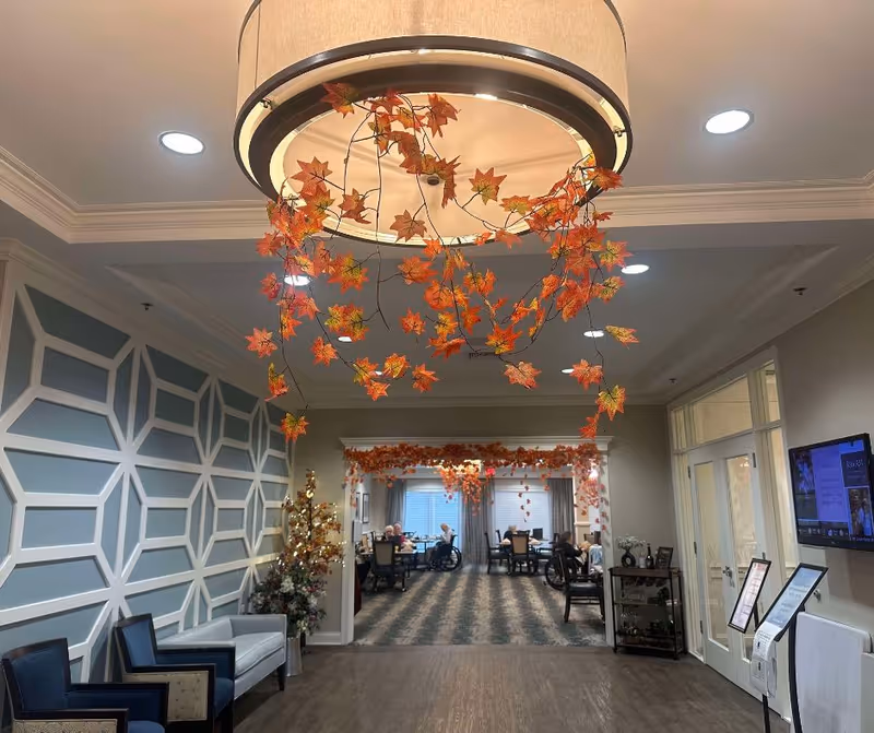 Interior view of a senior living facility with a decorative ceiling light adorned with autumn leaves. The room has patterned carpet and several tables where elderly residents are seated, some in wheelchairs. The walls feature geometric paneling and there are chairs and a small table with flowers near the entrance.