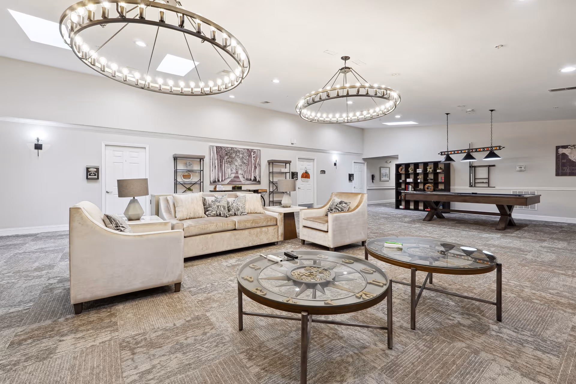 A spacious and well-lit common area with beige sofas and armchairs arranged around two round glass coffee tables. The room features two large circular chandeliers hanging from the ceiling, a pool table with overhead lighting, and shelves with decorative items along the back wall. The walls are painted light gray, and the floor is carpeted in a neutral tone.