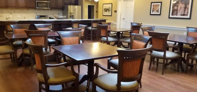 A dining area with multiple wooden tables and chairs arranged on a wooden floor. In the background, there is a kitchen area with a refrigerator, stove, and cabinets. The walls are decorated with framed pictures.