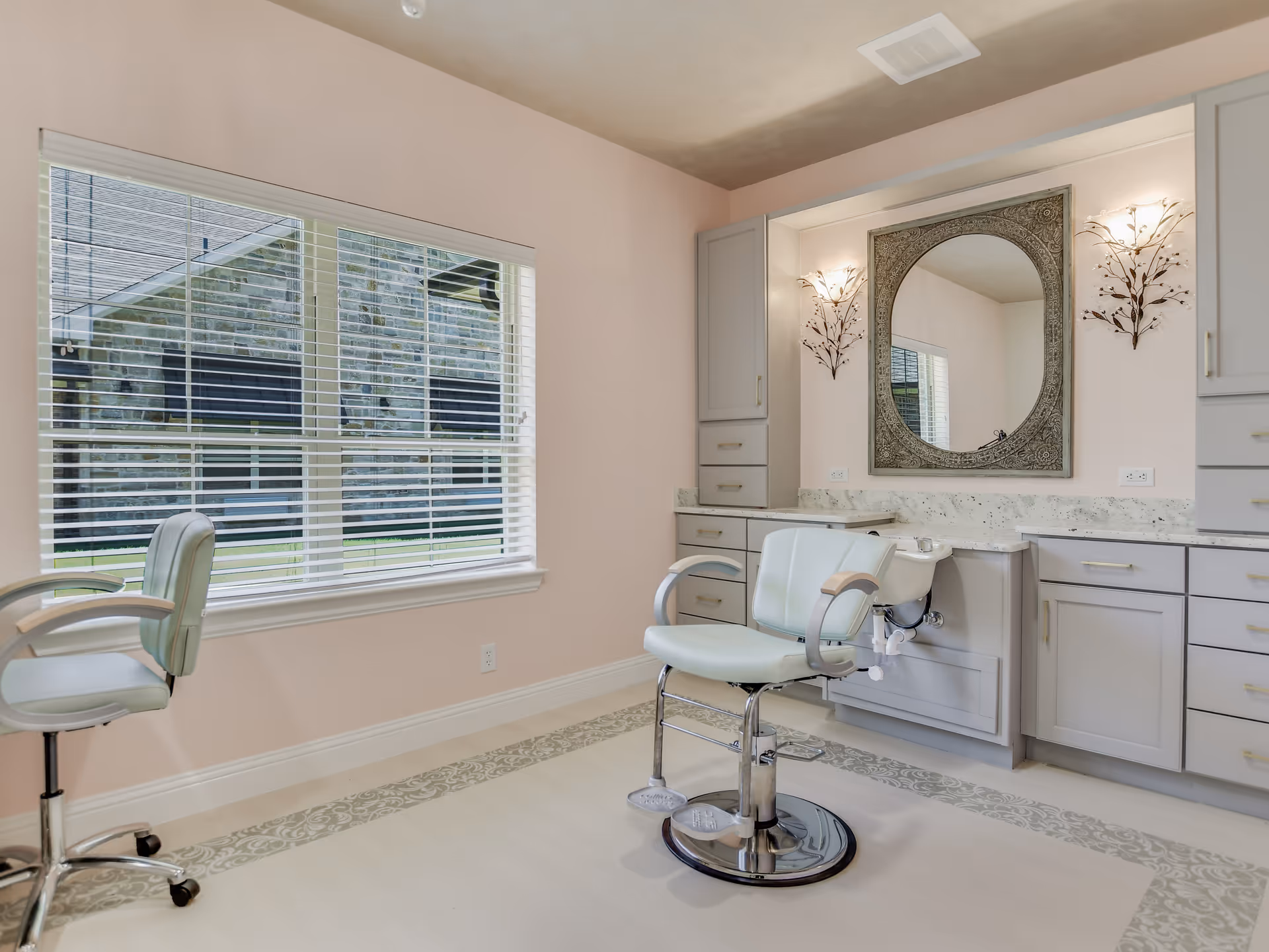 A bright and clean salon area with two light blue salon chairs, a large ornate oval mirror mounted on a wall with soft pink paint, and built-in cabinetry with marble countertops. A large window with white blinds lets in natural light, and decorative wall sconces flank the mirror.