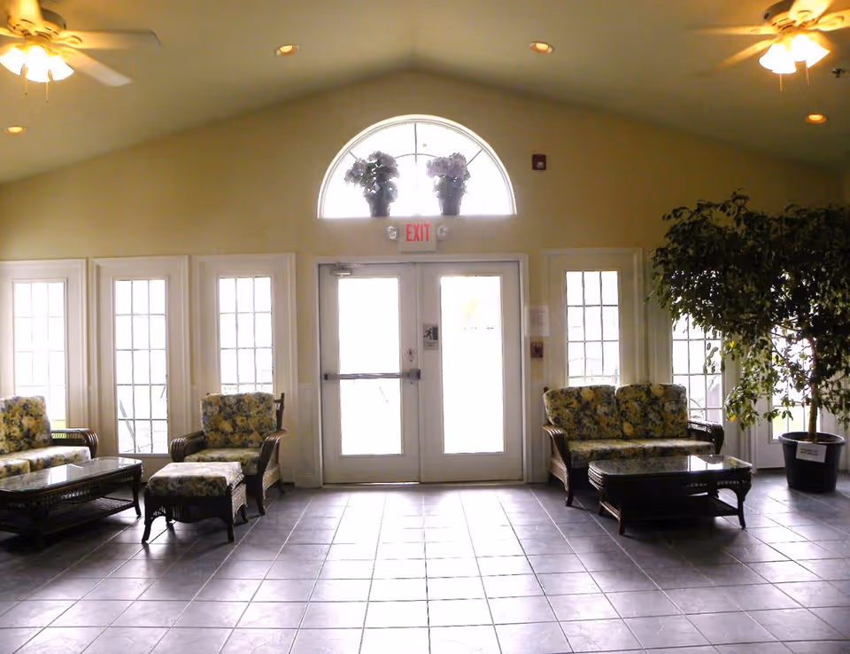 A bright interior common area with a tiled floor, two ceiling fans, and a set of double glass doors with an exit sign above. There are floral-patterned cushioned wicker chairs and sofas arranged around glass-top coffee tables on either side of the doors. A large potted plant is placed on the right side near the windows, and two flower pots are positioned on the window ledge above the doors.