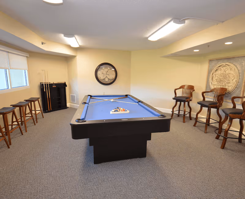 A recreational room with a blue felt pool table in the center, pool cues resting on the table, a rack with additional pool cues in the corner, four wooden bar stools with cushioned seats along the right wall, and a large round wall clock on the back wall. The room has beige walls, carpeted floor, and a window with a roller shade on the left side.