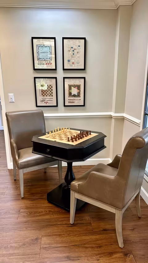 Two brown leather chairs positioned around a black octagonal table with a chessboard and chess pieces set up. The room has wooden flooring and four framed game-related prints hanging on a light-colored wall.
