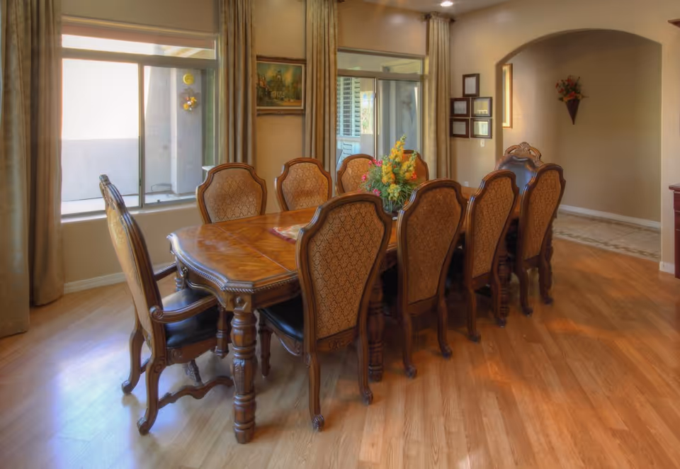 A dining room with a long wooden table surrounded by eight ornate chairs with patterned upholstery. The room has wooden flooring, two large windows with curtains, a floral centerpiece on the table, framed pictures on the wall, and a wall-mounted flower arrangement in the corner.
