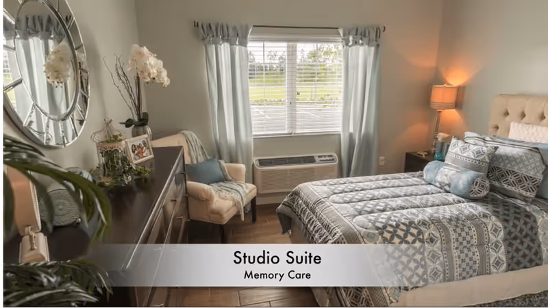 Furnished studio bedroom with a made bed, dresser, armchair, window and lamp.