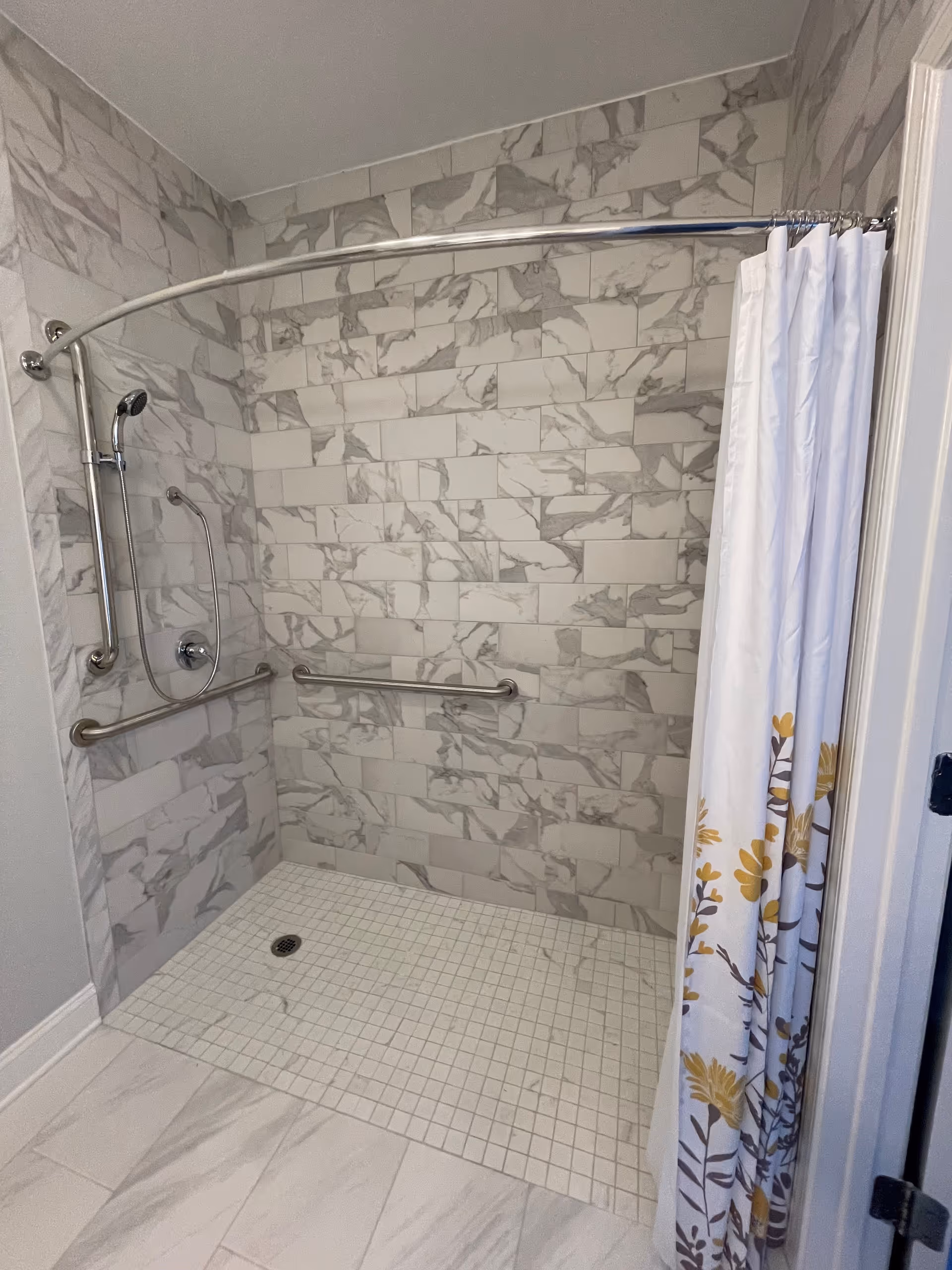 A walk-in shower with marble-patterned wall tiles and a tiled floor. The shower features a curved chrome shower curtain rod with a white curtain decorated with yellow and brown floral designs. There are two horizontal grab bars and a handheld showerhead attached to a vertical bar on the left wall.
