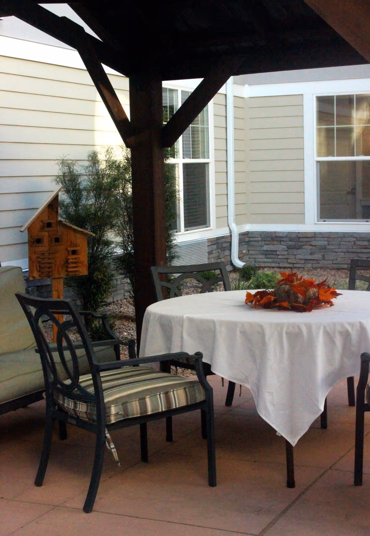 Outdoor patio area with a round table covered by a white tablecloth and decorated with autumn leaves centerpiece. Surrounding the table are metal chairs with striped cushions. The patio is partially covered by a wooden pergola, and the background shows beige siding walls with windows and some greenery.