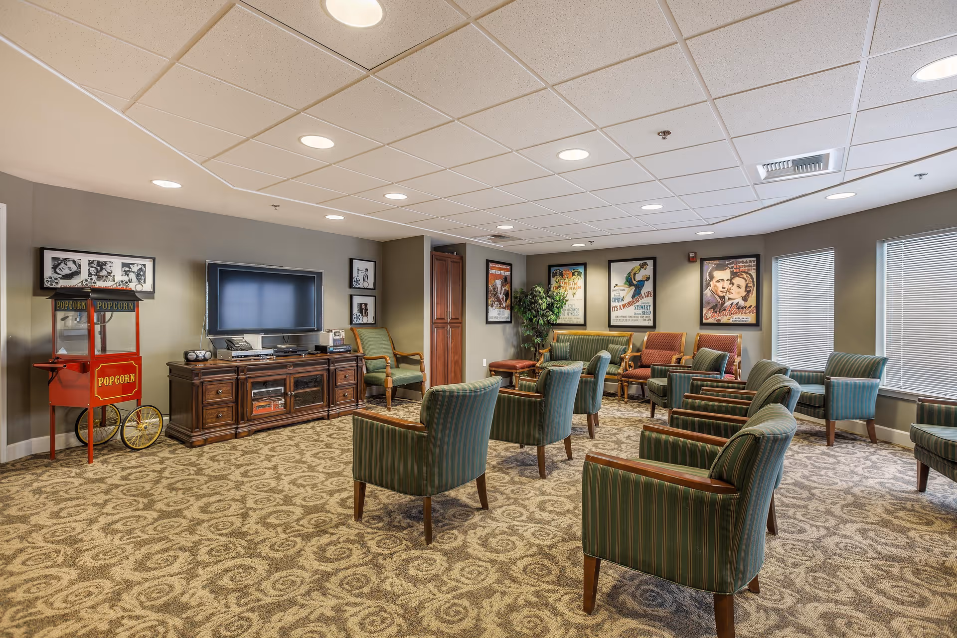 A spacious common living room area in Parkview Estates featuring multiple green striped armchairs arranged in rows facing a large flat-screen TV on a wooden media console. There is a vintage-style red popcorn machine on the left side and framed classic movie posters on the walls. The room has patterned carpet flooring, a drop ceiling with recessed lighting, and several windows with blinds allowing natural light.
