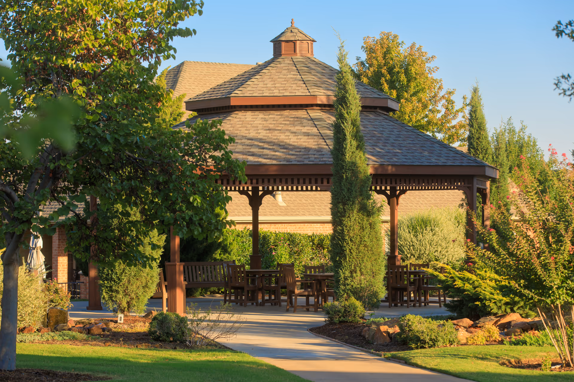 A wooden gazebo with a shingled roof surrounded by lush greenery and trees in a garden area. There are several wooden chairs and tables under the gazebo, with a paved walkway leading to it. The scene is bright and sunny with a clear blue sky.