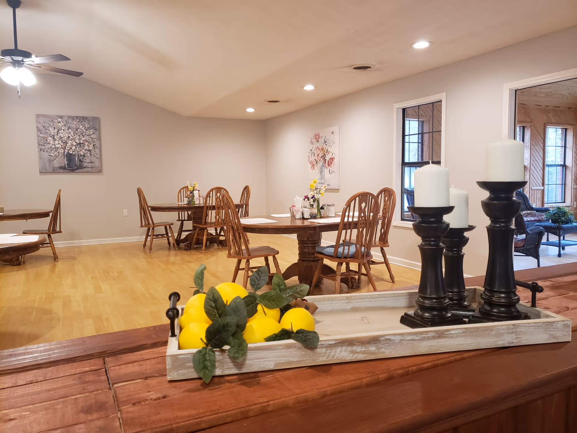 A dining room with wooden tables and chairs, decorated with small flower arrangements. In the foreground, there is a wooden tray holding artificial lemons and three black candle holders with white candles. The room has light-colored walls, a ceiling fan with lights, and a painting of flowers on the wall. There is a window and an open doorway leading to another room with wicker furniture and large windows.