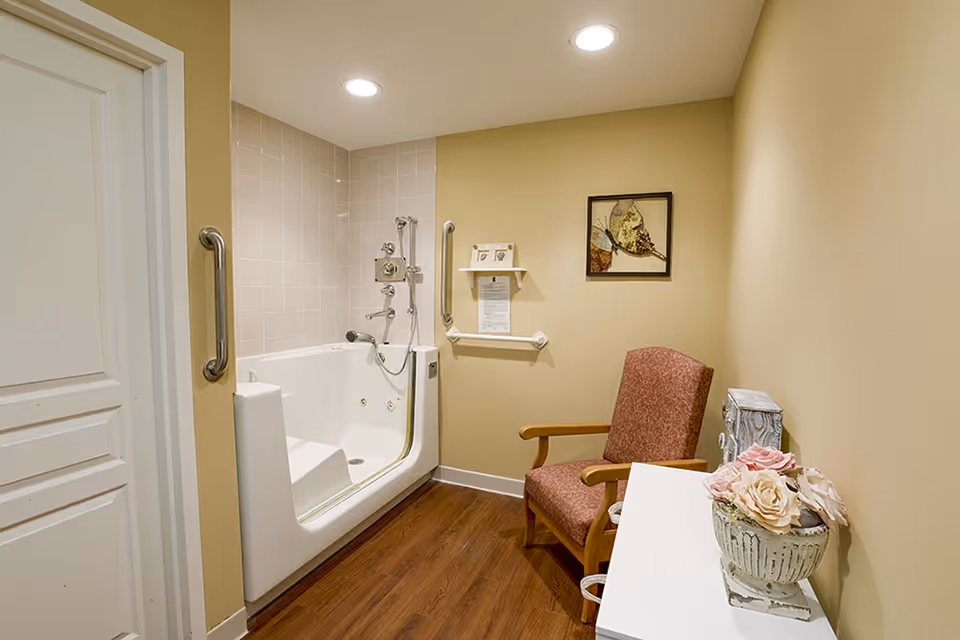A small bathing room with a walk-in bathtub featuring handrails and a handheld shower. The room has beige walls, wood flooring, a red upholstered armchair, a white cabinet with a decorative flower arrangement, and a framed butterfly picture on the wall.