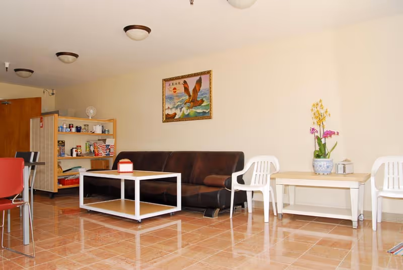 A living room area with a brown leather sofa, a white coffee table, two white plastic chairs, and a side table with a potted orchid plant. There is a painting of an eagle on the wall above the sofa and a shelving unit with various items in the background. The floor is tiled and the walls are light-colored.