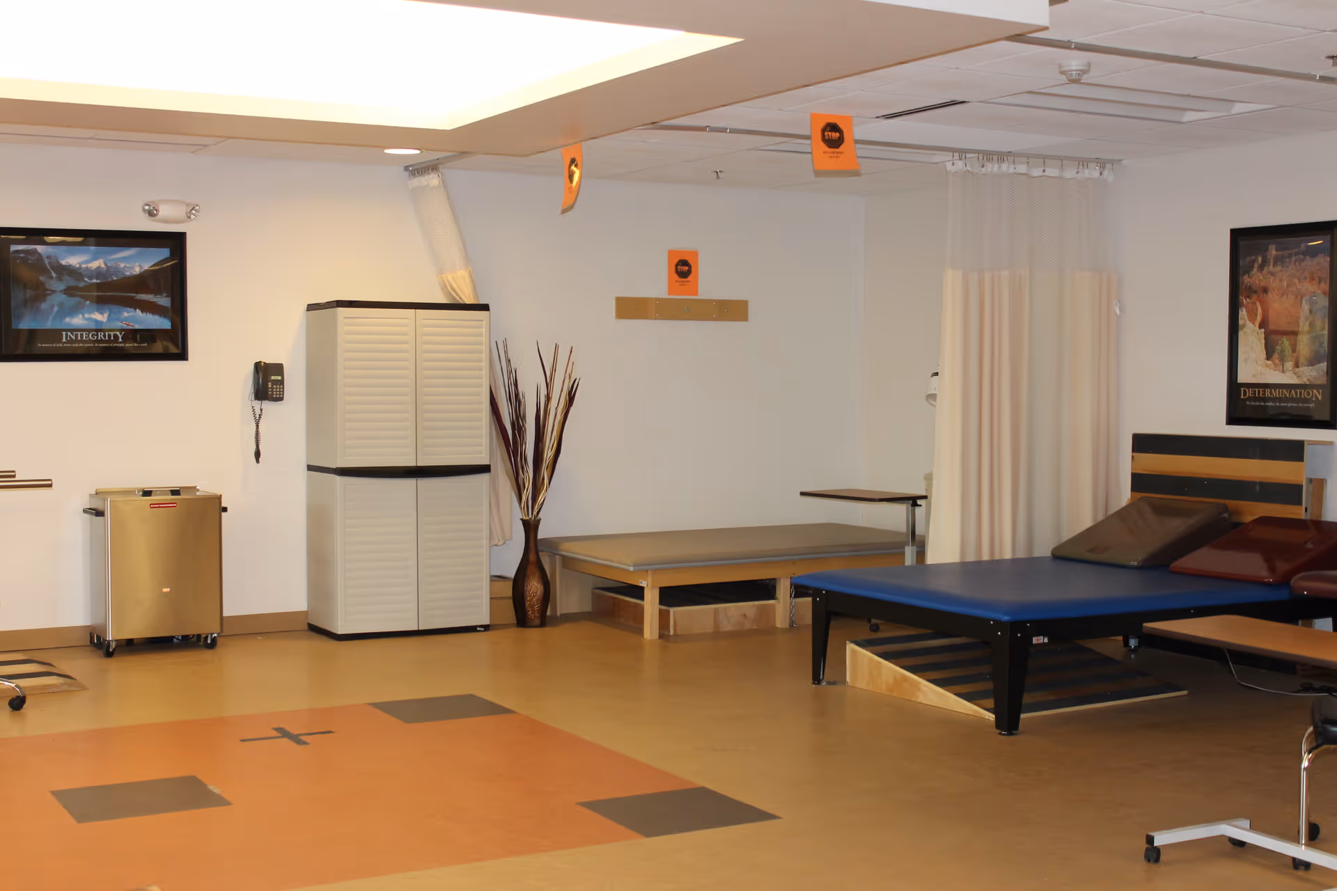 Rehabilitation therapy room containing treatment tables, storage cabinets, a privacy curtain, and framed posters on the walls.