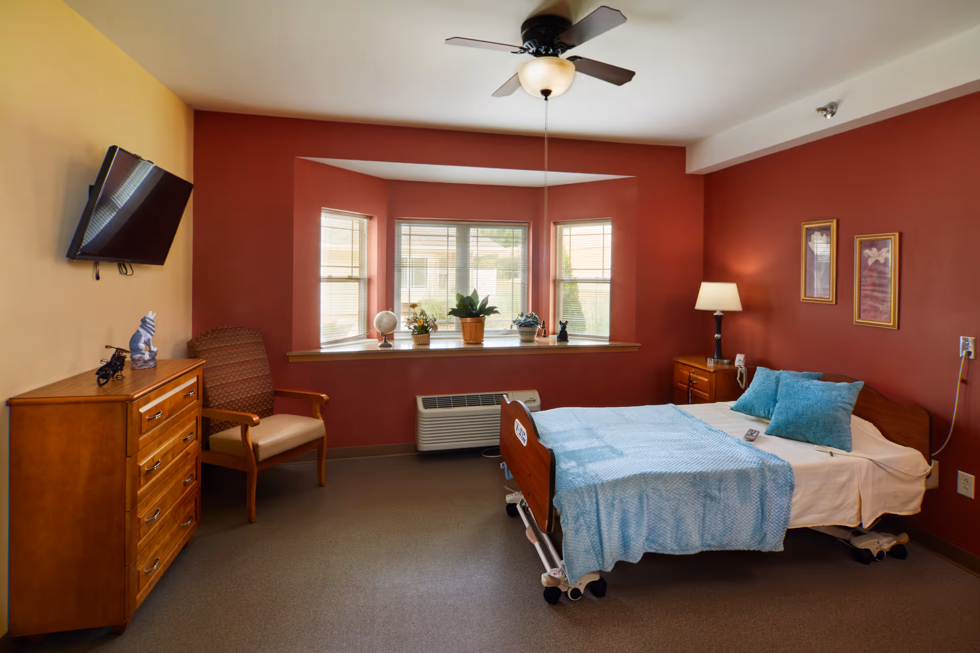 A cozy senior living bedroom with a hospital-style bed covered in light blue bedding and pillows. The room features a red accent wall with two framed pictures, a wooden nightstand with a lamp, a wooden dresser with decorative items, a comfortable armchair, a wall-mounted flat-screen TV, and a ceiling fan. A bay window with blinds and several potted plants lets in natural light.