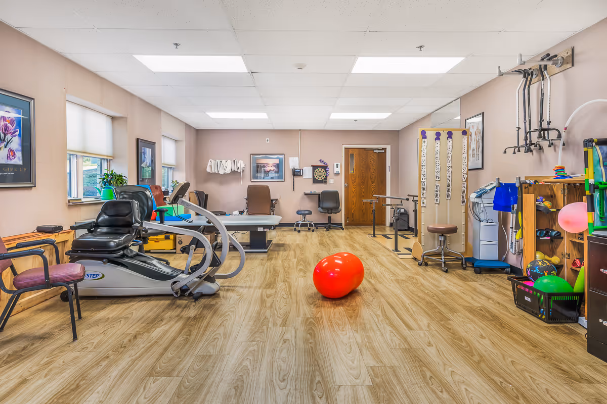 A spacious therapy or rehabilitation room with exercise equipment including a recumbent stepper, therapy chairs, parallel bars, and various therapy tools. The room has wood-patterned flooring, beige walls, fluorescent ceiling lights, and windows with blinds. There are framed pictures on the walls and a bright red exercise ball in the center of the room.