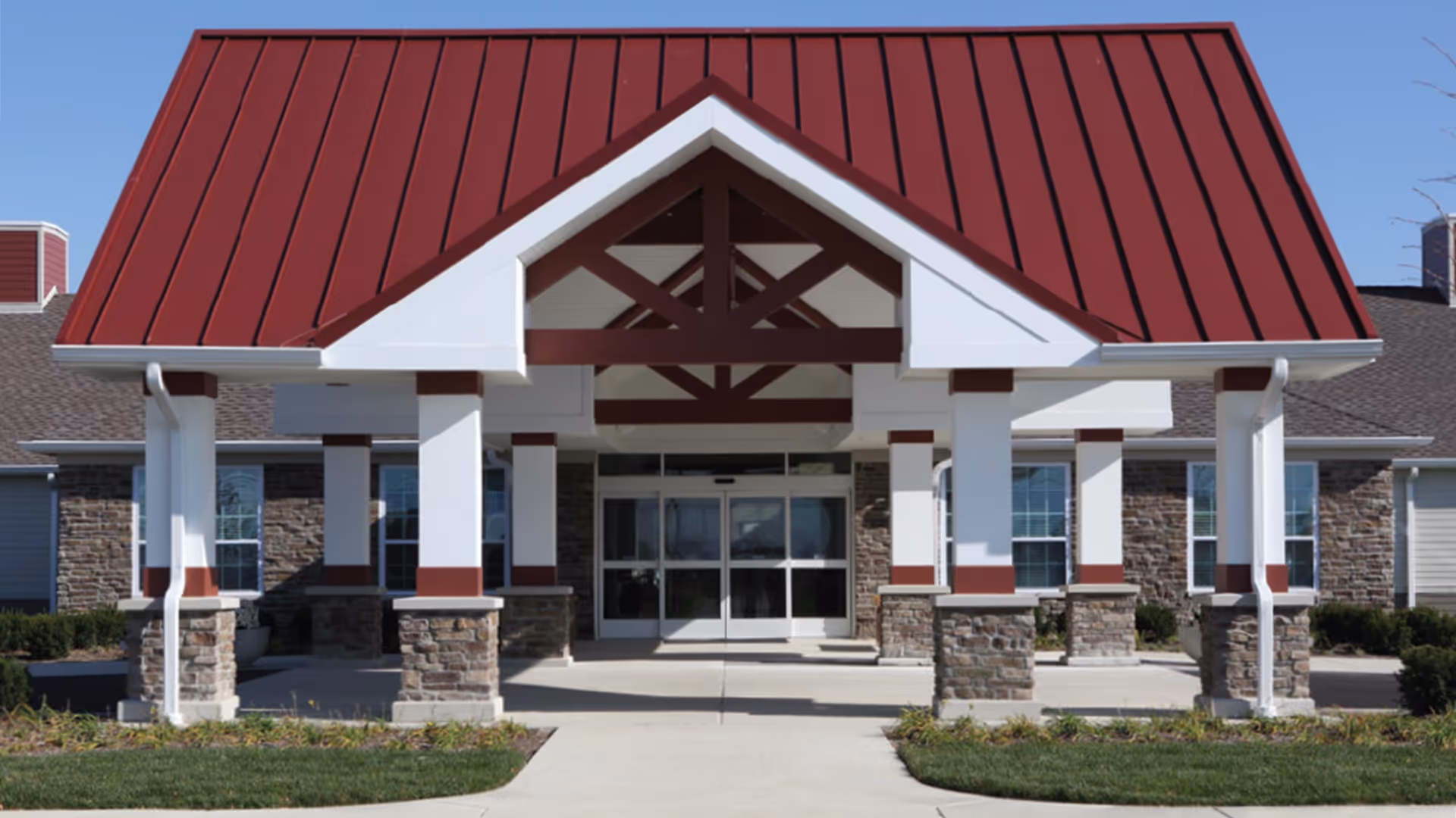 Front entrance of a senior living building with a red gabled metal roof, white columns with stone bases, and glass double doors.
