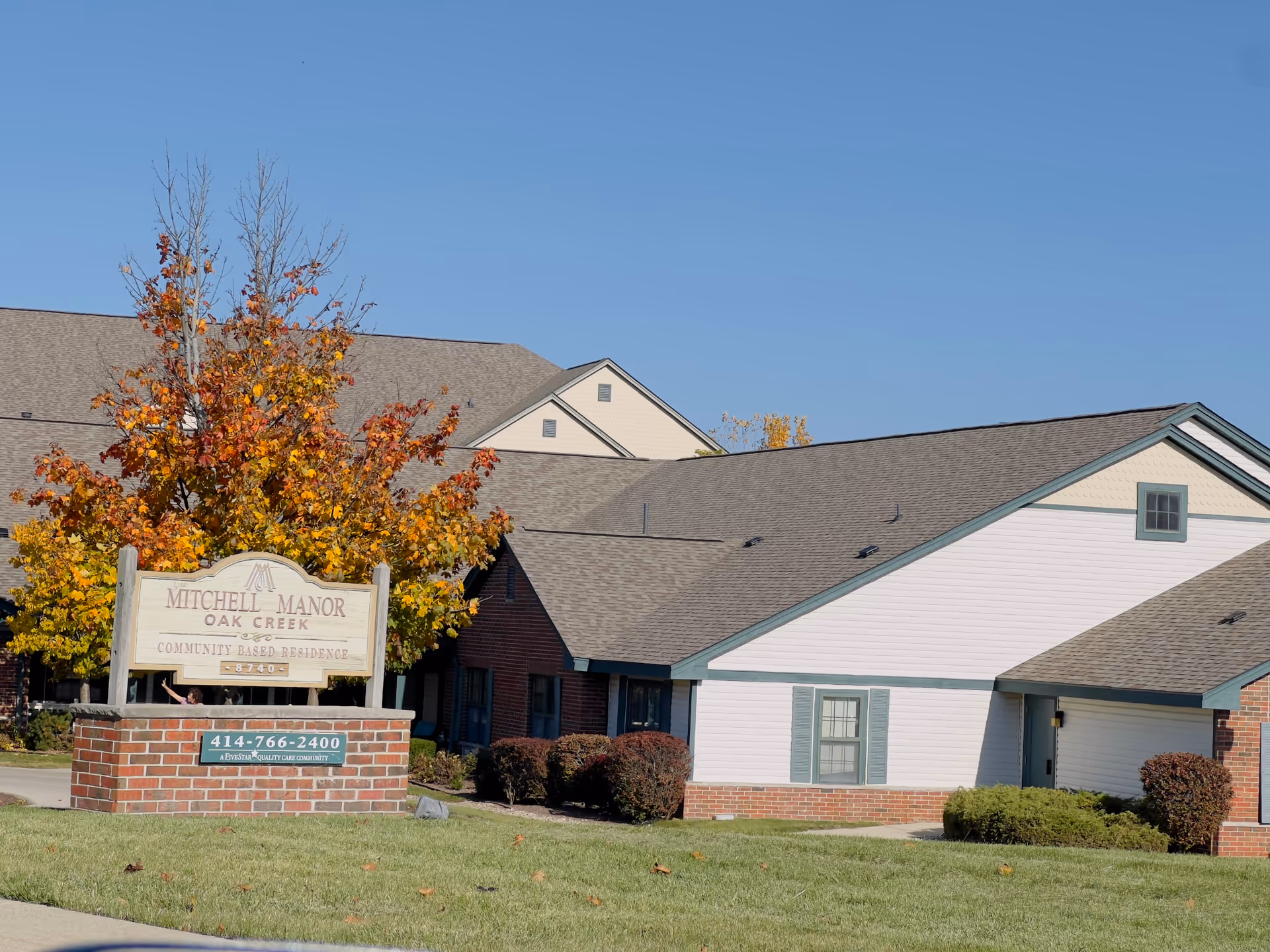 Exterior view of Mitchell Manor Oak Creek, a community-based residence. The building has a combination of brick and white siding with green trim around the windows. In front of the building is a sign with the facility's name and contact information, surrounded by a lawn and autumn-colored trees under a clear blue sky.