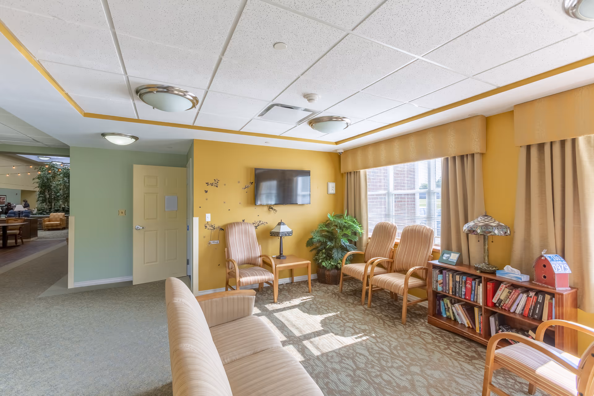 A bright and cozy common area with yellow walls and large windows letting in natural light. The room features several cushioned chairs arranged around a small wooden table with a lamp, a wall-mounted TV, a bookshelf filled with books, and a potted plant. The ceiling has recessed lighting and a drop ceiling design. There is an open doorway leading to another area with more seating and greenery.