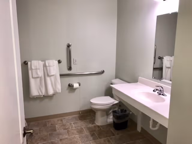 A clean and accessible bathroom with a white toilet, a white sink with a countertop, a large mirror above the sink, two white towels hanging on a towel rack, a grab bar next to the toilet, and a small trash bin under the sink. The floor is tiled with brown tiles.