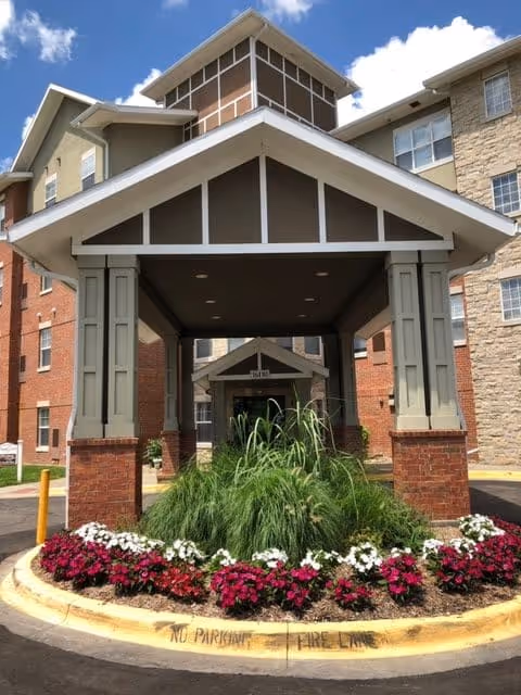 Entrance canopy of a senior living facility named The Gardens at Creekside, featuring a covered drop-off area with a landscaped circular flower bed containing green plants and red and white flowers. The building exterior is a mix of brick and stone with multiple windows under a blue sky with some clouds.